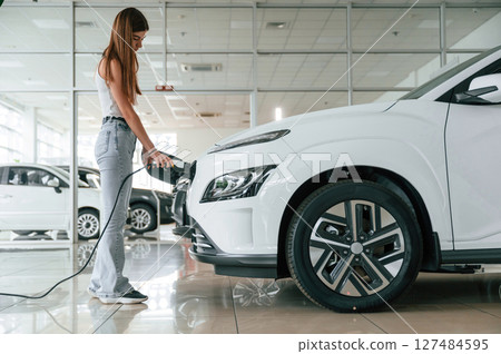 Side view. Charging the vehicle. Beautiful woman is indoors with the modern car Side view. Charging the vehicle. Beautiful woman is indoors with the modern car 127484595