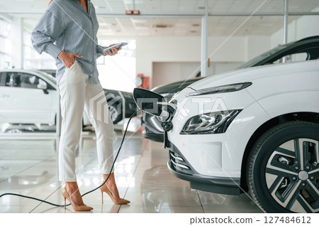 Standing, holding smartphone, automobile is charging. Close up view of woman that is with electric car 127484612