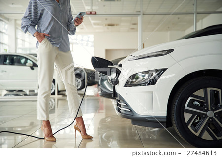 Standing, holding smartphone, automobile is charging. Close up view of woman that is with electric car 127484613