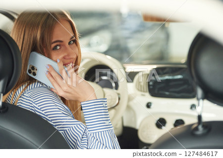 Sitting inside aand talking by phone. Beautiful woman is indoors with the modern car 127484617