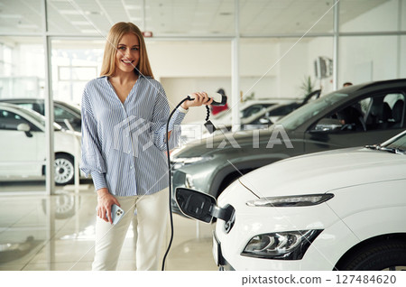 Electric car charger in hands. Beautiful woman is indoors Electric car charger in hands. Beautiful woman is indoors 127484620