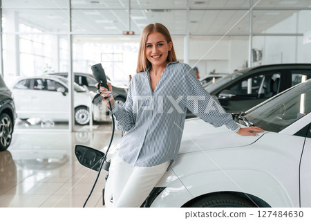 Leaning on the vehicle and holding charger for electric car. Beautiful woman is indoors 127484630