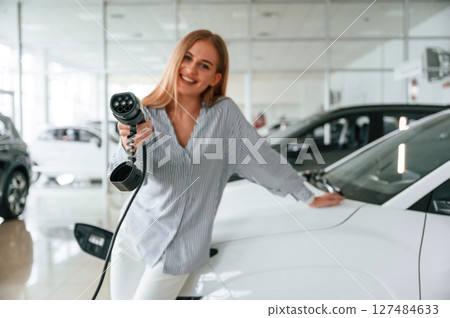 Leaning on the vehicle and holding charger for electric car. Beautiful woman is indoors 127484633