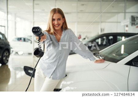 Leaning on the vehicle and holding charger for electric car. Beautiful woman is indoors 127484634