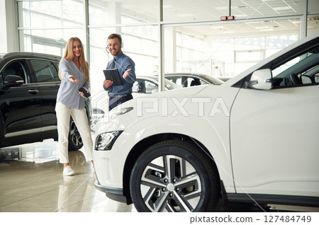 Beautiful white automobile. Man in formal clothes is with woman customer with the electric car Beautiful white automobile. Man in formal clothes is with woman customer with the electric car 127484749