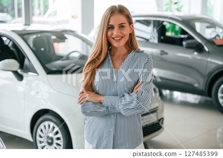 Standing with arms crossed. Beautiful woman is indoors with the modern car Standing with arms crossed. Beautiful woman is indoors with the modern car 127485399