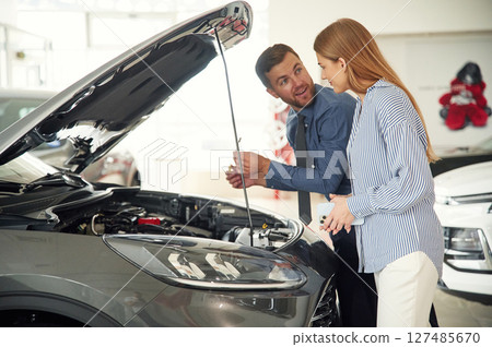 Under the hood. Man in formal clothes is with woman customer with the electric car 127485670