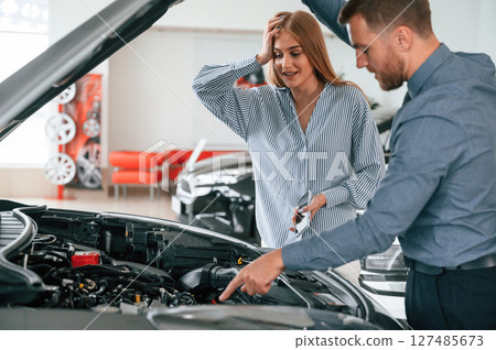Under the hood. Man in formal clothes is with woman customer with the electric car 127485673