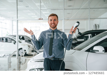 Smiling, holding the charger. Handsome car dealership worker is with electric vehicle 127485681