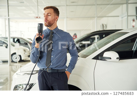 Having fun with charger. Handsome car dealership worker is with electric vehicle Having fun with charger. Handsome car dealership worker is with electric vehicle 127485683
