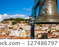 Close-up of historic bell in selective focus overlooking Lisbon, Portugal, colorful rooftops, iconic St Jorge castle, clear blue sky and some clouds in blurred background. City heritage, vibrant 127486797