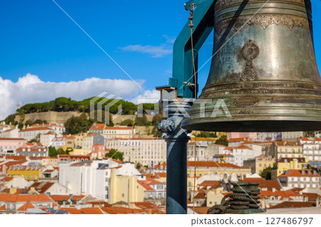 Close-up of historic bell in selective focus overlooking Lisbon, Portugal, colorful rooftops, iconic St Jorge castle, clear blue sky and some clouds in blurred background. City heritage, vibrant 127486797