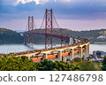 Iconic 25 de Abril bridge in Lisbon, Portugal, in heavy traffic, full with vehicles in transit at sunset. Red suspension bridge over Tagus River, major city landmark, transport route, lush greenery 127486798