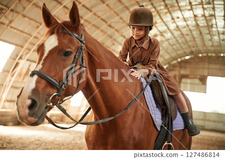 Riding the animal. Cute little girl is with horse indoors Riding the animal. Cute little girl is with horse indoors 127486814