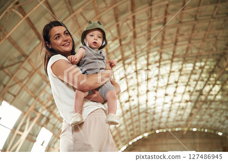Holding kid in hands. Gorgeous woman with her little baby son is with horse indoors Holding kid in hands. Gorgeous woman with her little baby son is with horse indoors 127486945