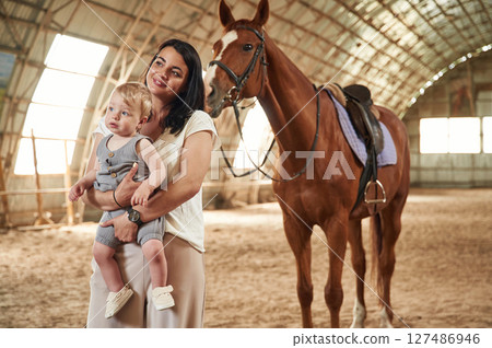 Holding kid in hands. Gorgeous woman with her little baby son is with horse indoors Holding kid in hands. Gorgeous woman with her little baby son is with horse indoors 127486946