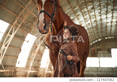 In jockey clothes. Cute little girl is with horse indoors In jockey clothes. Cute little girl is with horse indoors 127487062