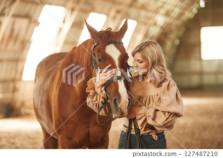 Embracing the head of the animal. Beautiful young woman is with horse indoors 127487210