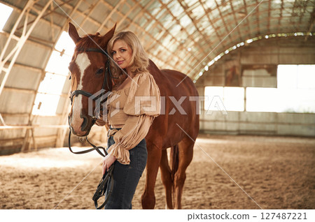 Sunny day. Standing with animal. Beautiful young woman is with horse indoors 127487221