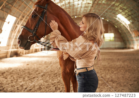 Sunny day. Standing with animal. Beautiful young woman is with horse indoors 127487223