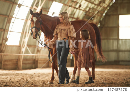 Woman with her little daughter is with horse indoors Woman with her little daughter is with horse indoors 127487278