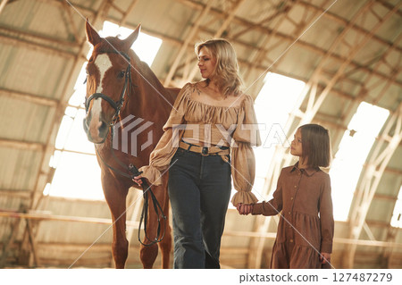 Woman with her little daughter is with horse indoors Woman with her little daughter is with horse indoors 127487279