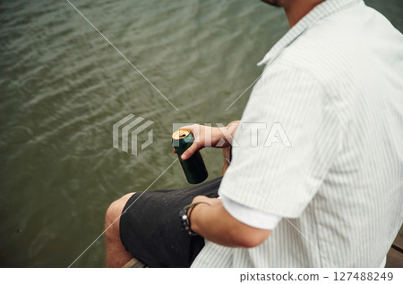 Close up view of man that is sitting by the water with beer in hands Close up view of man that is sitting by the water with beer in hands 127488249