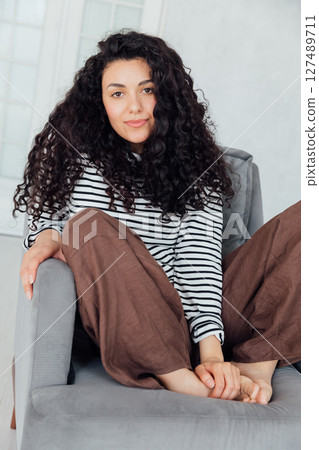 Curly brunette woman in coat sitting on chair and smiling Curly brunette woman in coat sitting on chair and smiling 127489711