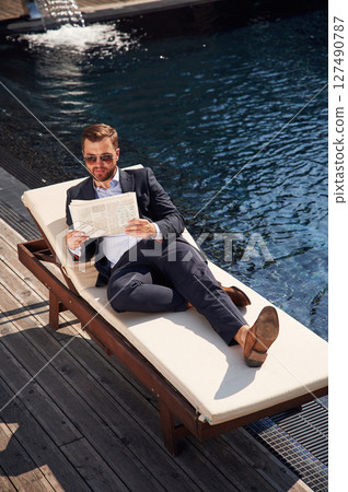 Holding newspaper. Handsome man in formal clothes is in the pool 127490787