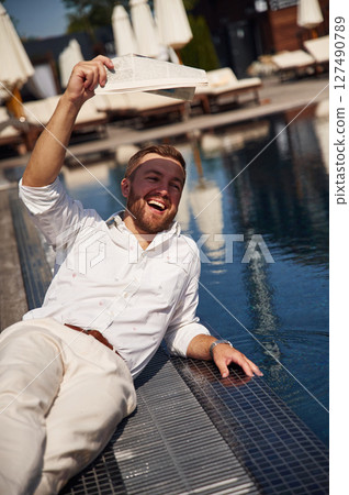 Covering himself by newspaper from bright sunlight. Handsome man in formal clothes is in the pool 127490789