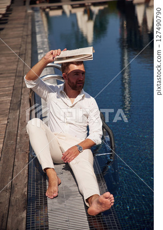 Covering himself by newspaper from bright sunlight. Handsome man in formal clothes is in the pool 127490790