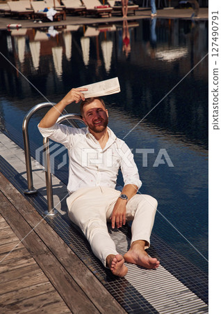 Covering himself by newspaper from bright sunlight. Handsome man in formal clothes is in the pool Covering himself by newspaper from bright sunlight. Handsome man in formal clothes is in the pool 127490791