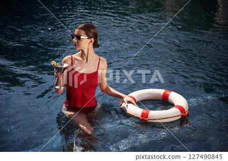 Standing and holding cocktail. Young woman in red swimsuit is in the pool 127490845
