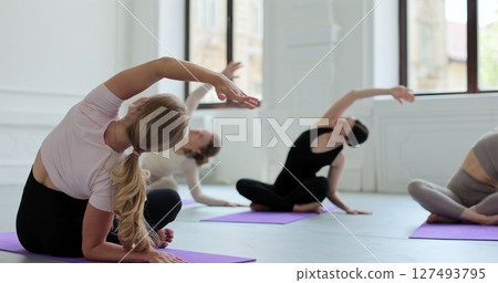 Women repeating asanas after female yoga coach, sitting on mats in bright studio with big windows. Concept of healthy lifestyles and self development. Women repeating asanas after female yoga coach, sitting on mats in bright studio with big windows. Concept of healthy lifestyles and self development. 127493795