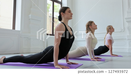 Group of four young women are practicing yoga on mats in a large, bright room. Concept of healthy lifestyles and self development. Group of four young women are practicing yoga on mats in a large, bright room. Concept of healthy lifestyles and self development. 127493972