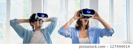 Couple experiencing virtual reality through headsets in bright modern living room 127494014