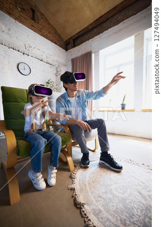 Father and child exploring virtual reality with VR headsets in bright modern living room setting Father and child exploring virtual reality with VR headsets in bright modern living room setting 127494049