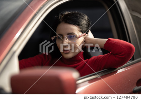 Sitting in the driver's seat of a car, a beautiful girl with glasses wears a red sweater and gazes thoughtfully outside, enjoying a warm sunny day and reflecting on her surroundings 127494790