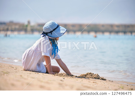 Pretty child girl in big hat and white dress playing with wet sand on background of blue sky and clear ocean lagoon water Pretty child girl in big hat and white dress playing with wet sand on background of blue sky and clear ocean lagoon water 127496475