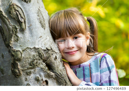 Portrait of pretty child girl leaning to a tree trunk in autumn park relaxing. Cute female kid enjoying warm fall weather in forest. 127496489