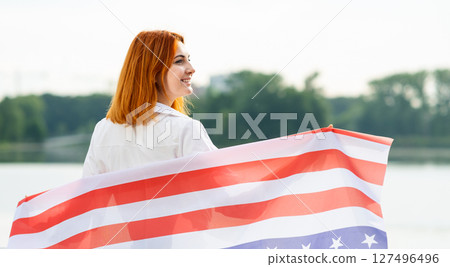 Portrait of happy smiling red haired girl with USA national flag in her hands. Pretty young woman celebrating United States independence day. 127496496