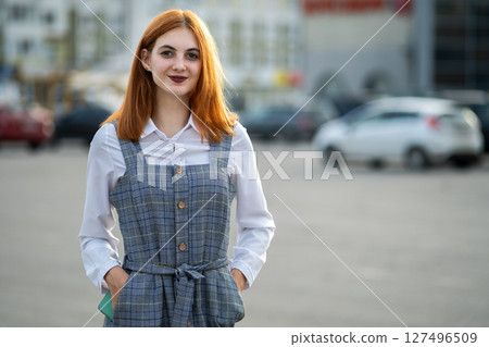 Portrait of a smiling teenage girl with red hair and clear eyes wearing fashionable casual dress standing outside in warm summer day. 127496509