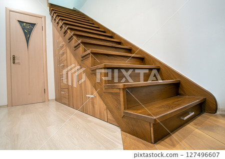 Modern architecture interior with luxury hallway with glossy wooden stairs in multi-storey house. Custom built pullout cabinets on glides in slots under stairs. Use of space for storage. Modern architecture interior with luxury hallway with glossy wooden stairs in multi-storey house. Custom built pullout cabinets on glides in slots under stairs. Use of space for storage. 127496607