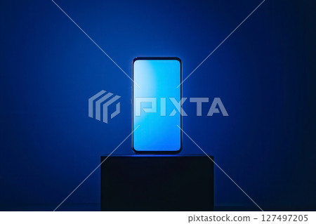 Illuminated smartphone on a pedestal against a deep blue backdrop showcasing modern technology and design Illuminated smartphone on a pedestal against a deep blue backdrop showcasing modern technology and design 127497205
