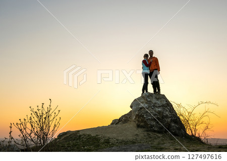 Man and woman hikers standing on a big stone at sunset in mountains. Couple together on a high rock in evening nature. Tourism, traveling and healthy lifestyle concept. Man and woman hikers standing on a big stone at sunset in mountains. Couple together on a high rock in evening nature. Tourism, traveling and healthy lifestyle concept. 127497616