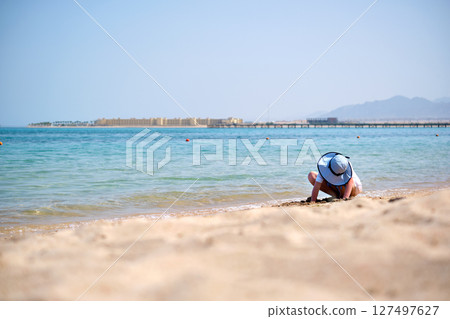 Lonely child girl playing with white sand on sea beach on background of blue sky, clear water and distant shore. Summer tourism and recreation concept Lonely child girl playing with white sand on sea beach on background of blue sky, clear water and distant shore. Summer tourism and recreation concept 127497627