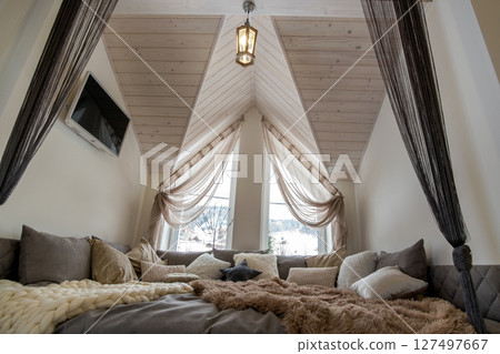 Interior of a modern house spacious hallway with big soft resting place. Contemporary wide sofa with many pillows and light window onder wooden loft ceiling. 127497667