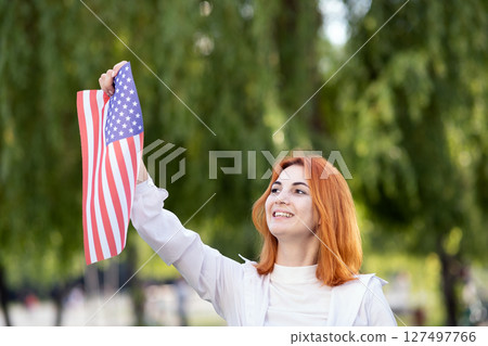 Happy young red haired woman holding USA national flag in her hands outdoors in summer. Positive girl celebrating United States independence day. 127497766