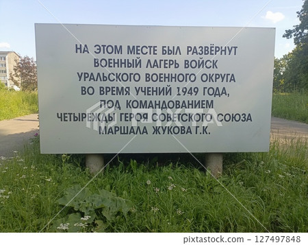 A sign in Russian detailing the history of a military camp established in 1949 under the command of a Soviet hero. Surrounded by greenery and grass. A sign in Russian detailing the history of a military camp established in 1949 under the command of a Soviet hero. Surrounded by greenery and grass. 127497848