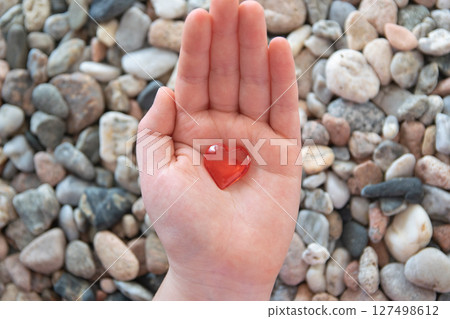 Red bright glass heart in child hand on the stones background. Concept of Valentine's day and romantic love. Red bright glass heart in child hand on the stones background. Concept of Valentine's day and romantic love. 127498612